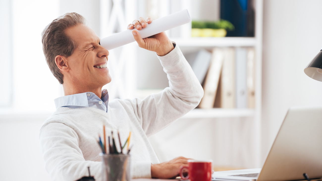Looking for new opportunities. Playful mature man holding handmade paper telescope and looking through it with smile while sitting at his working place